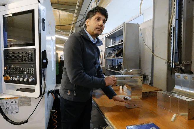 Ingénieur d’ACM France tenant une pièce métallique, debout dans un atelier à côté d’une machine industrielle.