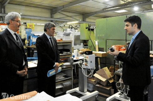 Deux représentants officiels en costume assistent à une présentation technique par un ingénieur dans un atelier industriel rempli d’équipements et d’outils.